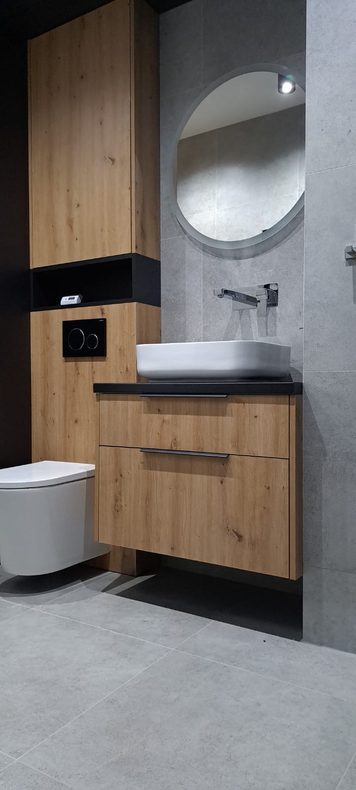 Nowoczesna łazienka z drewnianą szafką pod umywalkę z dwoma szufladami i czarnym blatem, okrągłe lustro z podświetleniem LED, wisząca toaleta, szare płytki na ścianach i podłodze.