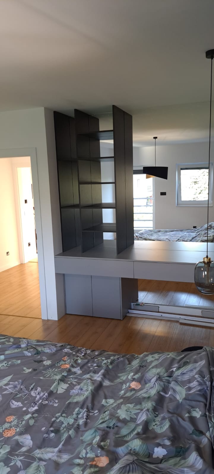 Szara zabudowa meblowa z półkami i szafkami, pełniąca funkcję ścianki działowej w sypialni, z widocznym łóżkiem w tle i drewnianą podłogą.