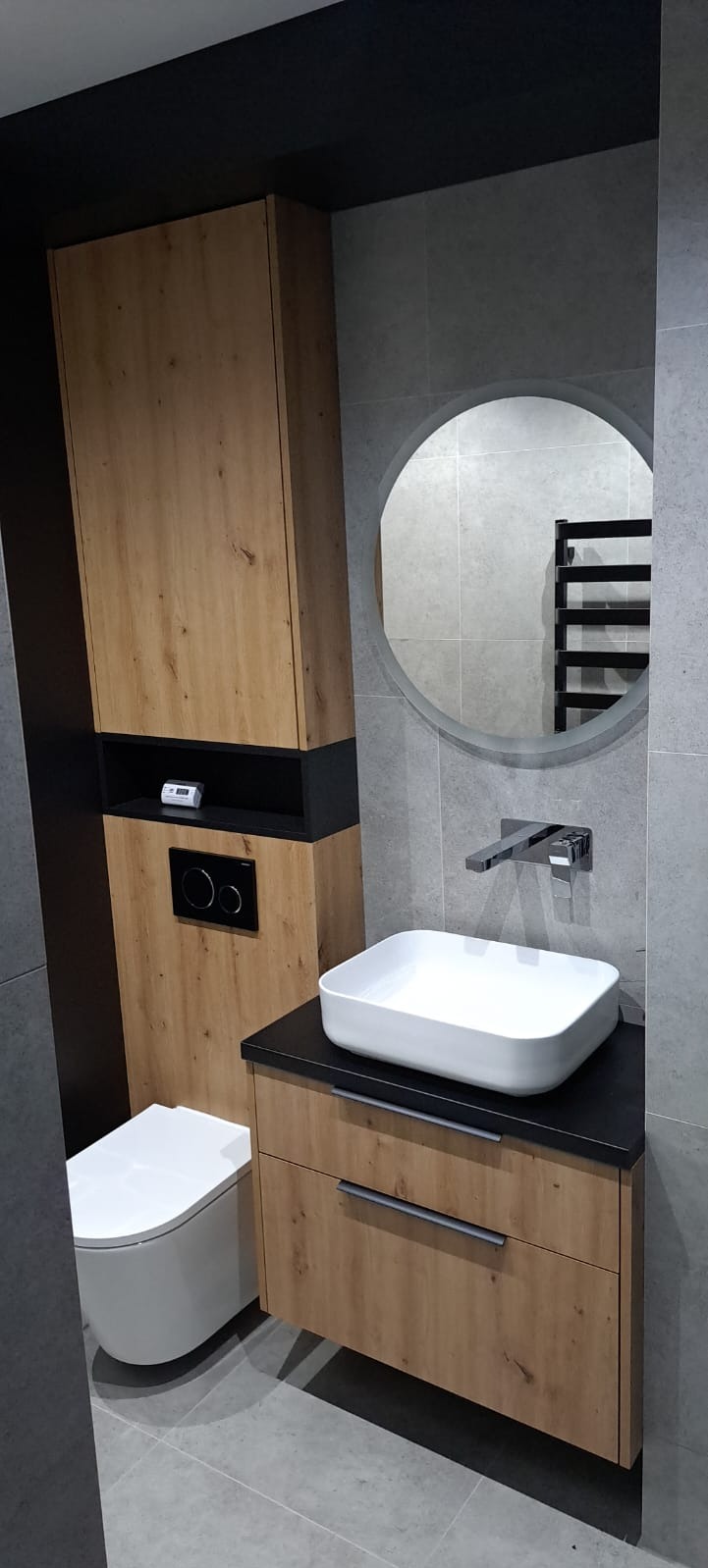 Nowoczesna łazienka z drewnianą szafką pod umywalką, wiszącą szafką z czarnymi akcentami i okrągłym lustrem na szarej płytce ściennej.