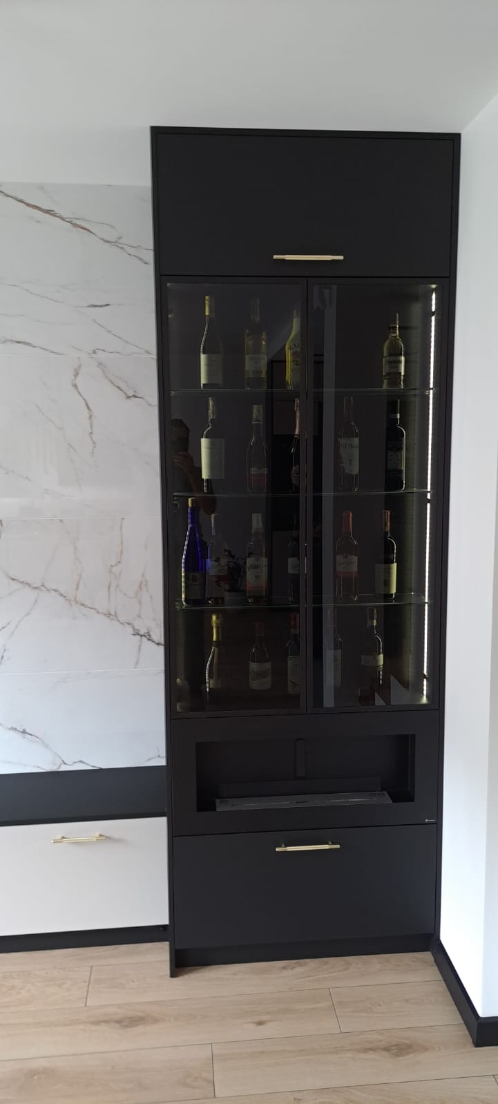 Czarna, wysoka szafa z przeszklonymi drzwiami, podświetlona od wewnątrz, wypełniona butelkami alkoholu, z złotymi uchwytami, umieszczona obok ściany z płytek imitujących marmur.