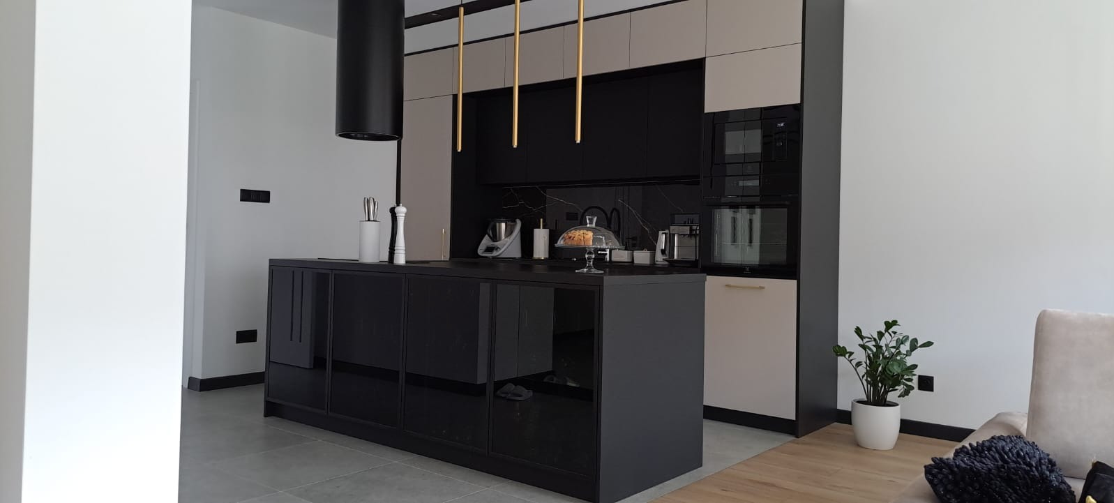 Nowoczesna kuchnia z czarną wyspą z połyskiem, marmurowym backsplashiem i złotymi lampami wiszącymi, uzupełniona o sprzęt AGD w zabudowie i dekoracyjne akcesoria.