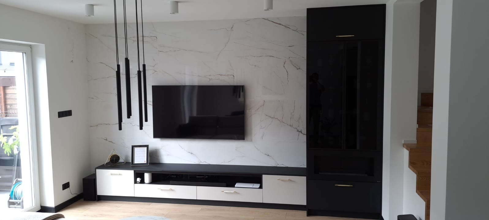 Nowoczesny salon z czarno-białymi meblami na wymiar, marmurową ścianą i wiszącymi lampami tubami, z widokiem na drewniane schody w tle.