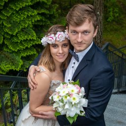 Foto-Mira-Żak - Uśmiechnięta para młoda w objęciach na schodach, panna młoda w wianku z białych i różowych kwiatów, pan młody w granatowym garniturze i muszce w kropki, trzymająca bukiet białych lilii i różowych...