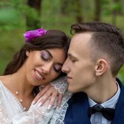 Foto-Mira-Żak - Uśmiechnięta brunetka z fioletowym kwiatem we włosach, opierająca głowę na dłoni, obok mężczyzna w granatowym garniturze i muszce, oboje z zamkniętymi oczami na tle zieleni.