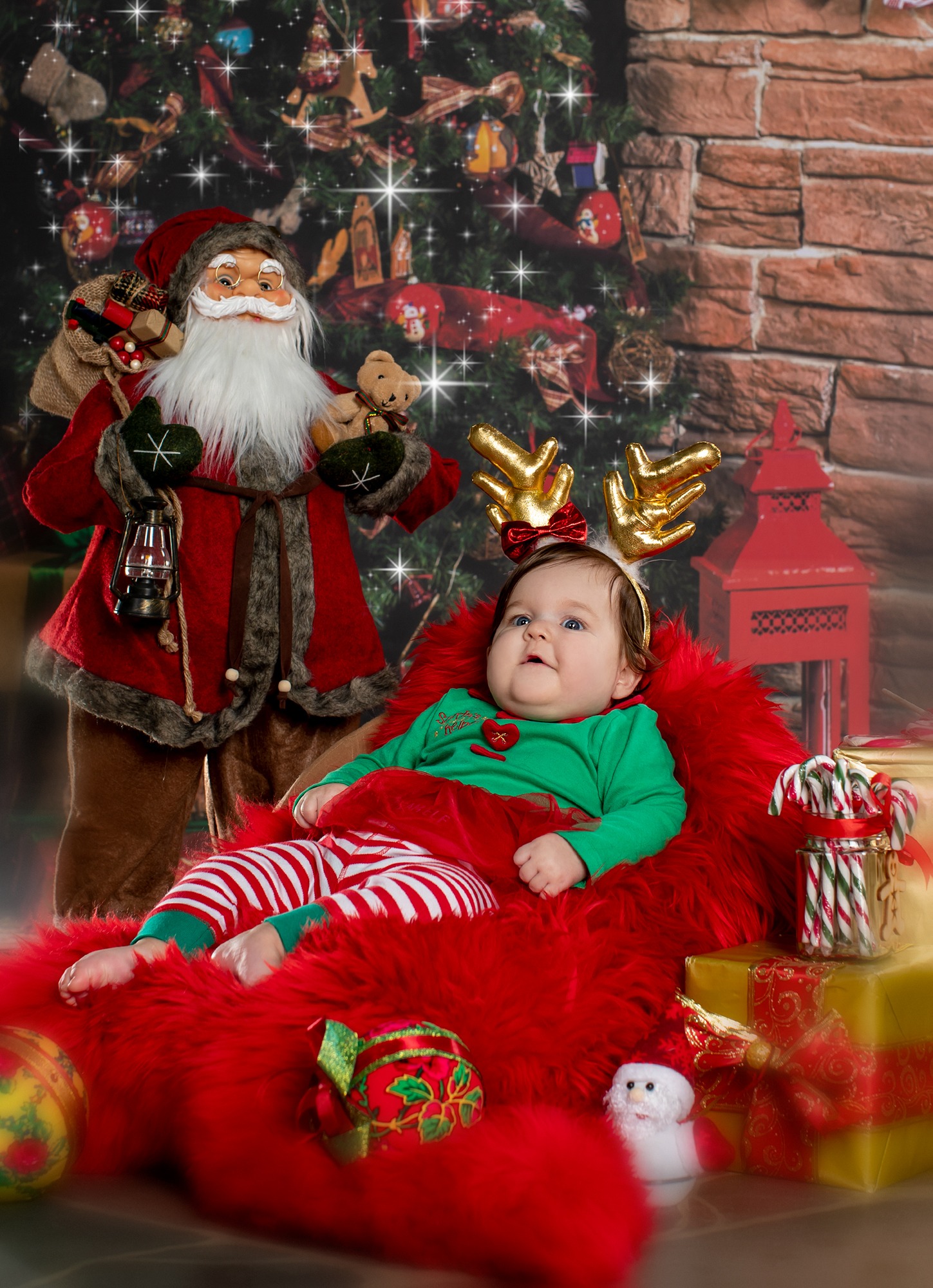 Uśmiechnięte niemowlę w świątecznym stroju z rogami renifera, siedzące na czerwonym futrze obok figurki Świętego Mikołaja i prezentów na tle choinki i kominka.