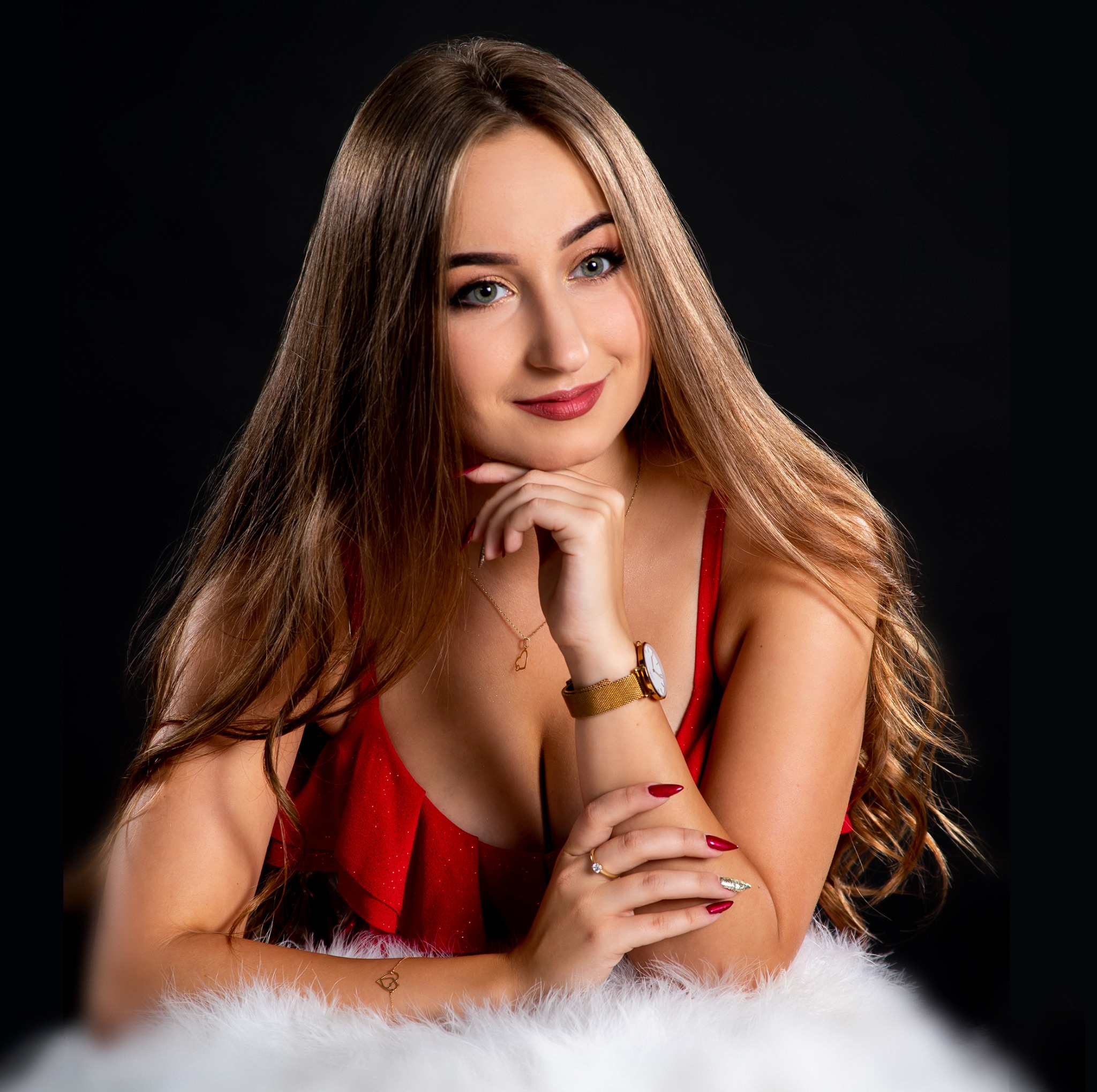 Portret młodej kobiety w czerwonej sukience, opierającej podbródek na dłoni, z widoczną biżuterią i manicure, na tle czerni.