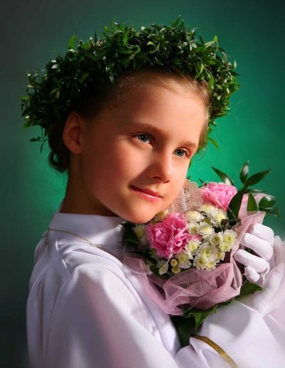 Dziewczynka w białej albie z zielonym wiankiem na głowie trzyma bukiet różowych i białych kwiatów na tle zielonego tła.
