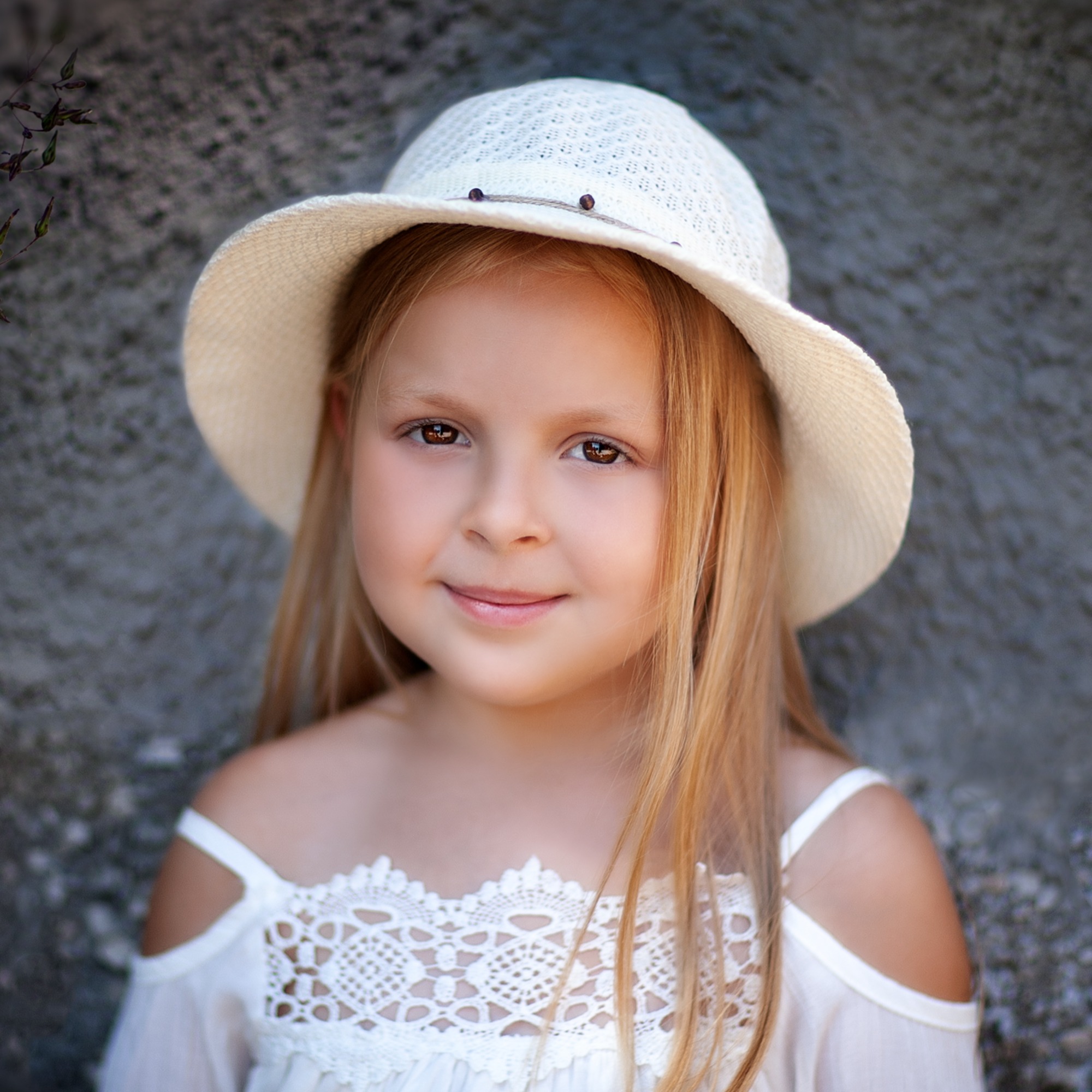 Portret uśmiechniętej dziewczynki w białym kapeluszu i koronkowej bluzce na tle szarej ściany.