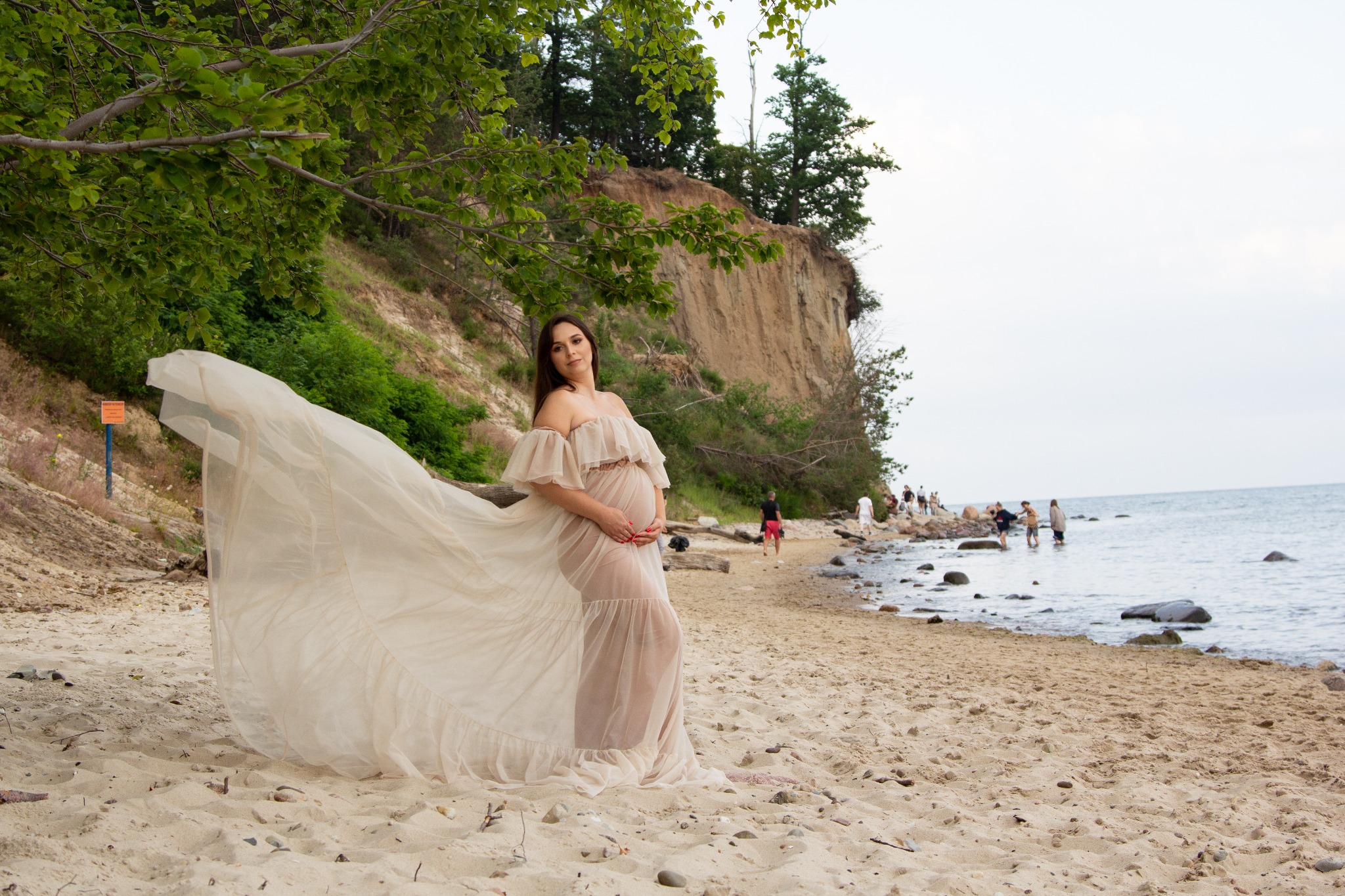 Kobieta w zwiewnej sukni ciążowej pozuje na piaszczystej plaży z klifem w tle, trzymając dłonie na brzuchu; na plaży w oddali widać spacerujących ludzi.