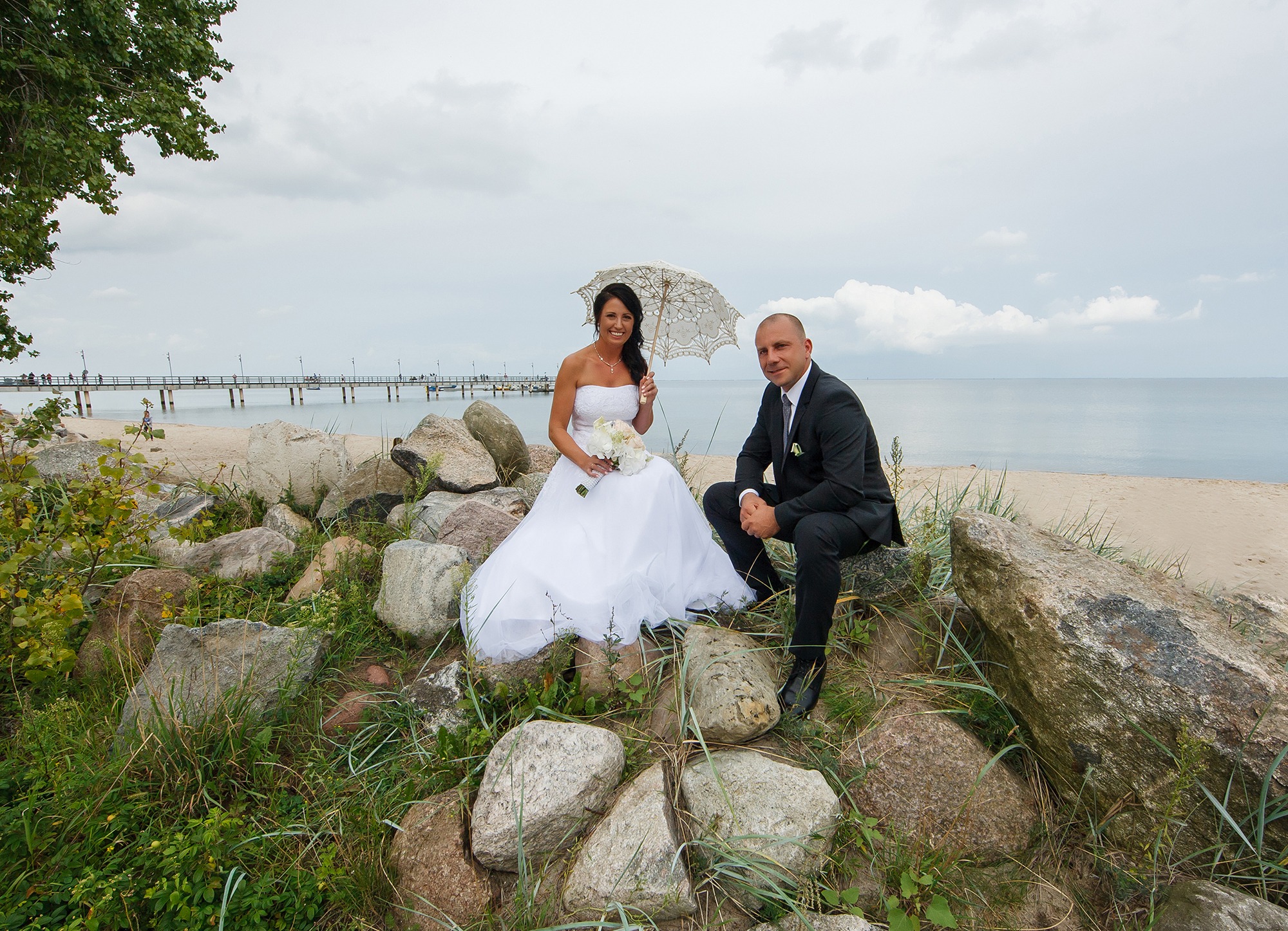 Para młoda siedzi na kamieniach z widokiem na morze w Rumi. Panna młoda trzyma bukiet i koronkowy parasol, pan młody w garniturze.