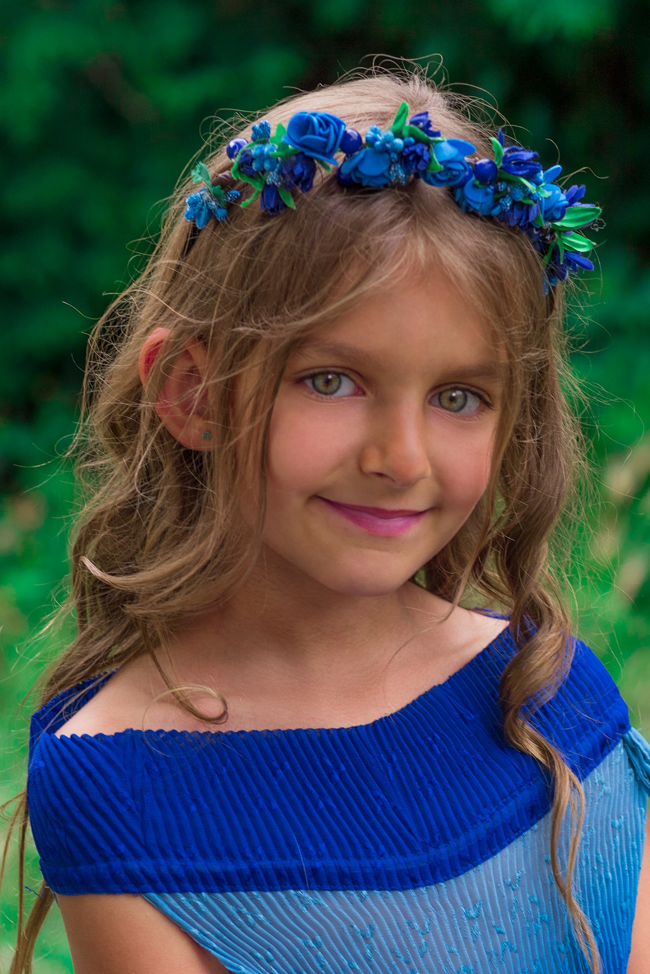 Portret uśmiechniętej dziewczynki z zielonymi oczami i wiankiem z błękitnych kwiatów na głowie, ubrana w plisowaną sukienkę w odcieniach błękitu, na rozmytym zielonym tle.