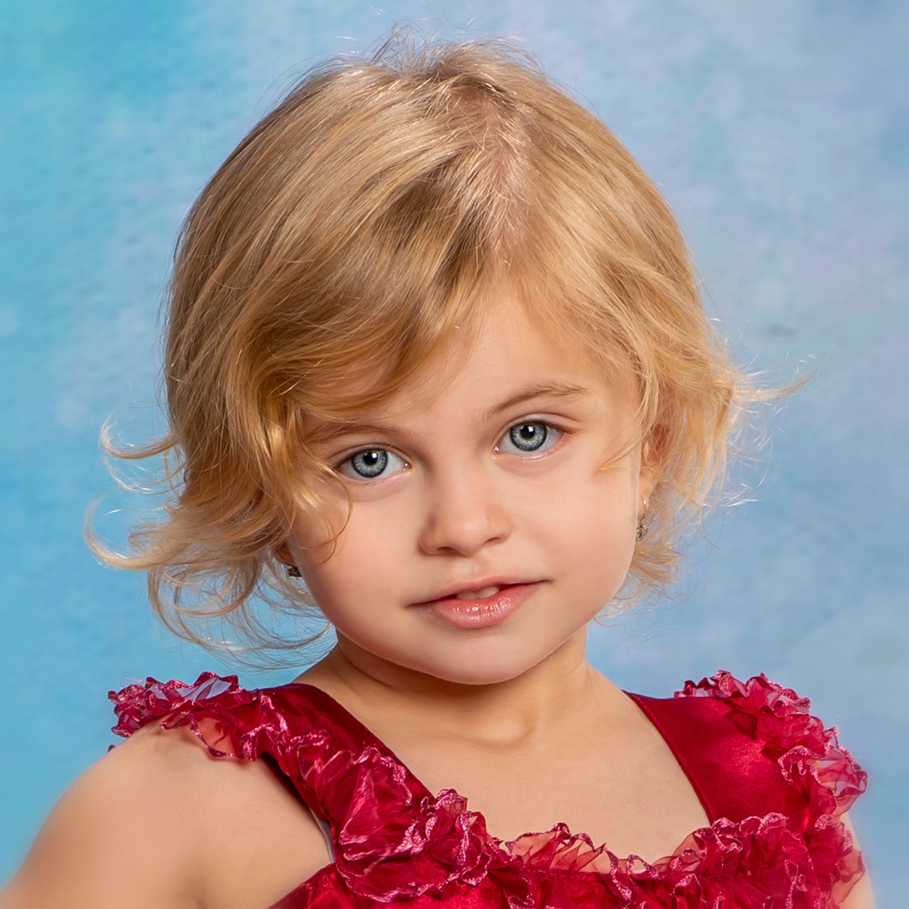 Portret uśmiechniętej dziewczynki z blond włosami i niebieskimi oczami w czerwonej sukience z falbankami na tle błękitnego tła.