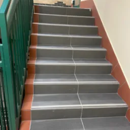 Schody wyłożone szarymi płytkami z pomarańczowymi akcentami i zieloną metalową barierką, widok z dołu.