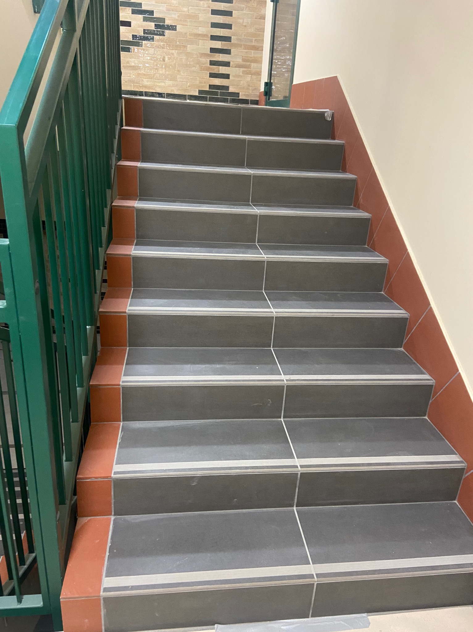 Schody wyłożone szarymi płytkami z pomarańczowymi akcentami i zieloną metalową barierką, widok z dołu.