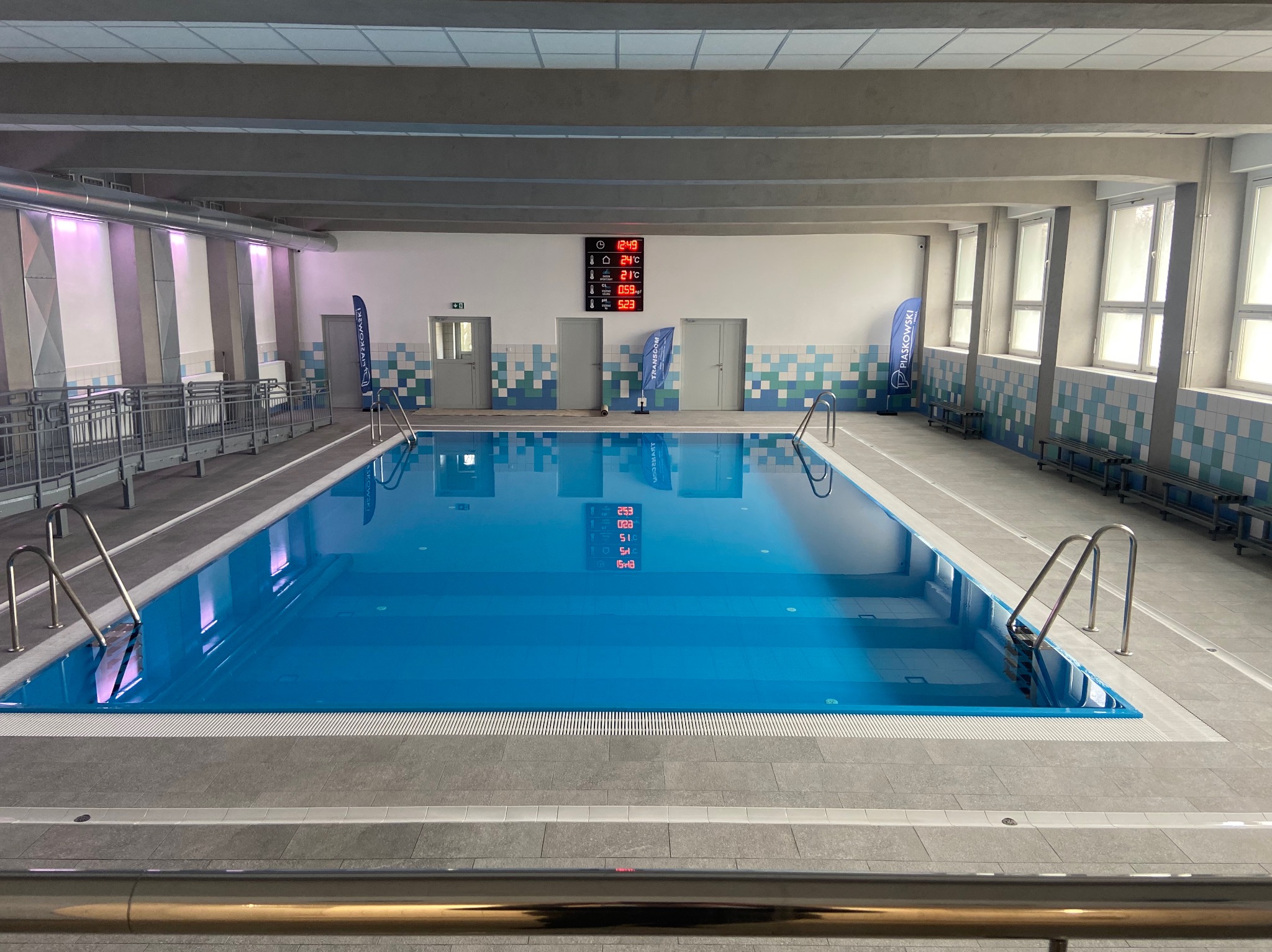 Nowoczesny basen kryty z błękitną wodą, szarymi płytkami, stalowymi drabinkami, cyfrowym wyświetlaczem parametrów wody i flagą firmową w tle.