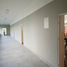 Daniel Nowik - Długi korytarz w budynku biurowym z szarymi ścianami, szarą podłogą z płytek i białymi drzwiami, widoczne oświetlenie sufitowe i biała skrzynka na ścianie.