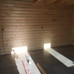 Daniel Nowik - Wnętrze w trakcie prac: drewniana boazeria na ścianach, szare płytki podłogowe z krzyżykami dystansowymi, widoczne kable elektryczne, poziomica i butelka wody.