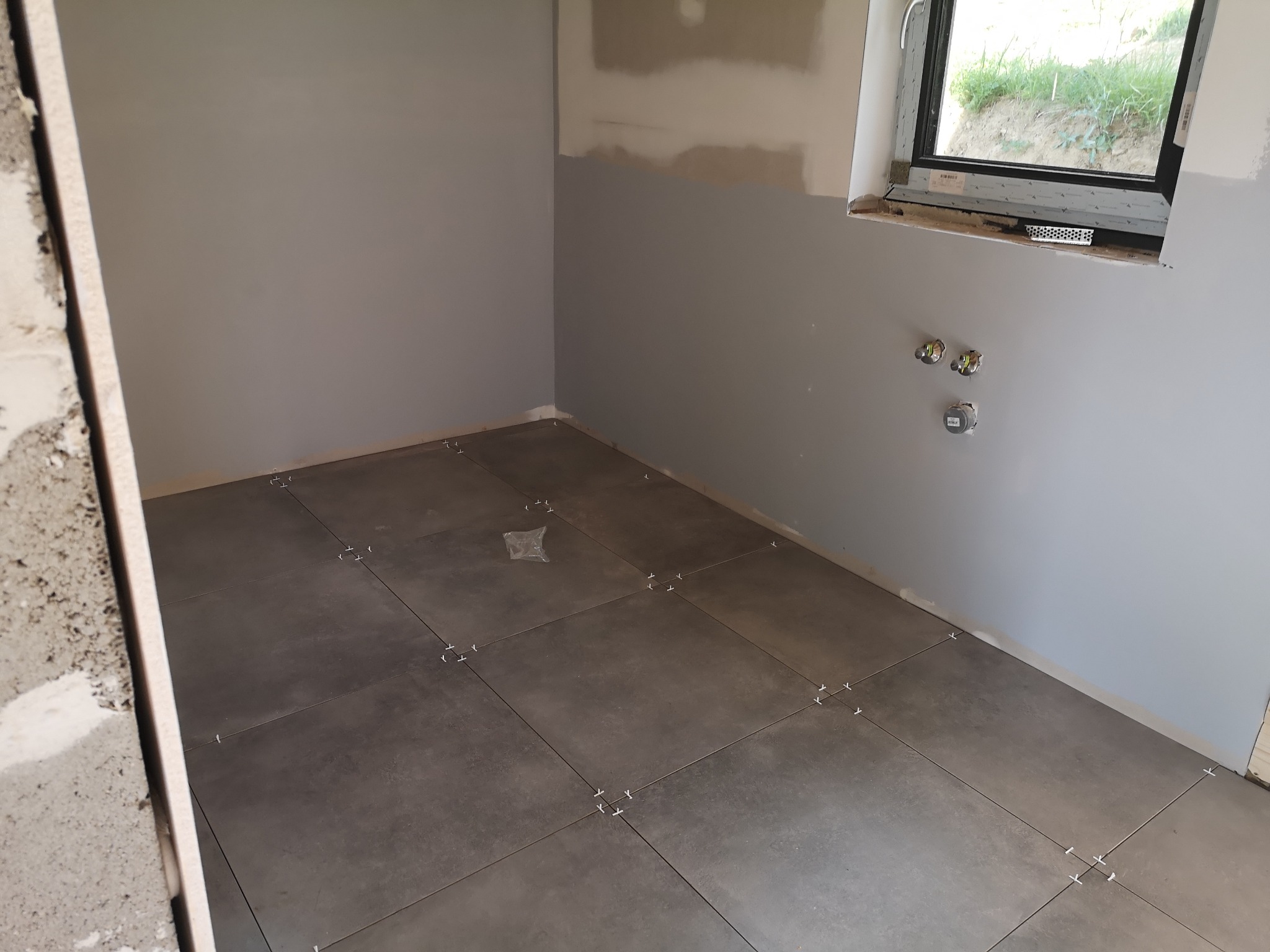 Układanie ciemnoszarych płytek podłogowych w pomieszczeniu w trakcie remontu, widoczne krzyżyki dystansowe, szare ściany częściowo zagruntowane, okno w tle.