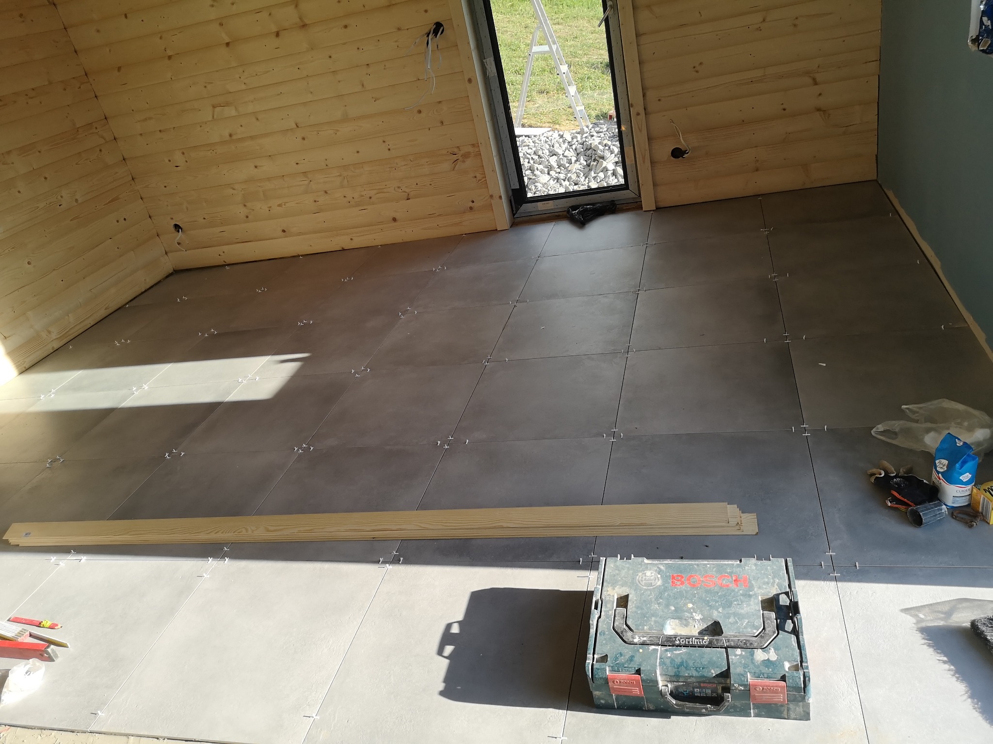 Układanie szarych płytek podłogowych w pomieszczeniu z drewnianą boazerią, widoczne krzyżyki dystansowe, skrzynka narzędziowa Bosch i deski przypodłogowe.