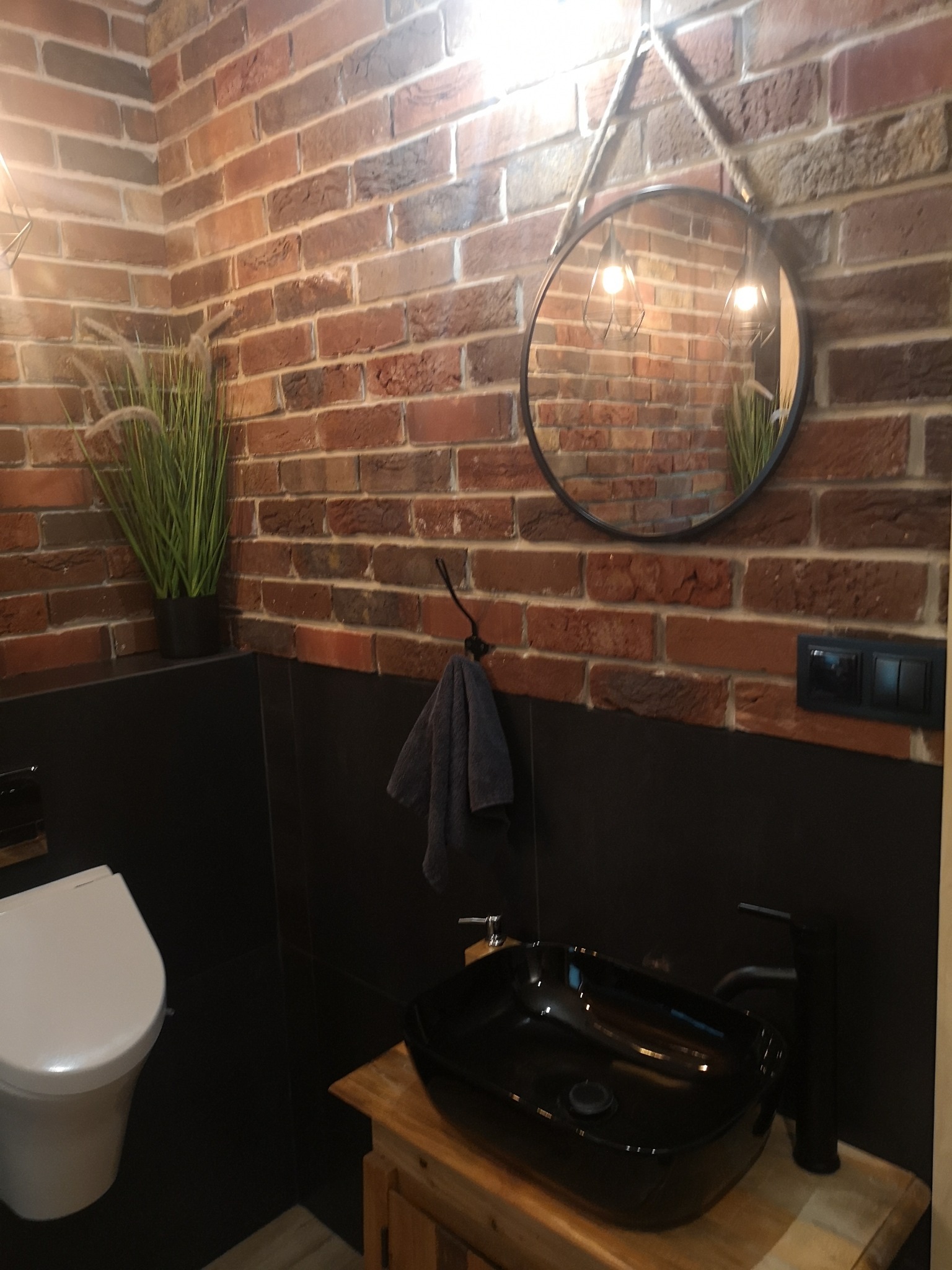 Nowoczesna łazienka z ceglaną ścianą, okrągłym lustrem na linie jutowej, czarną umywalką nablatową i toaletą.