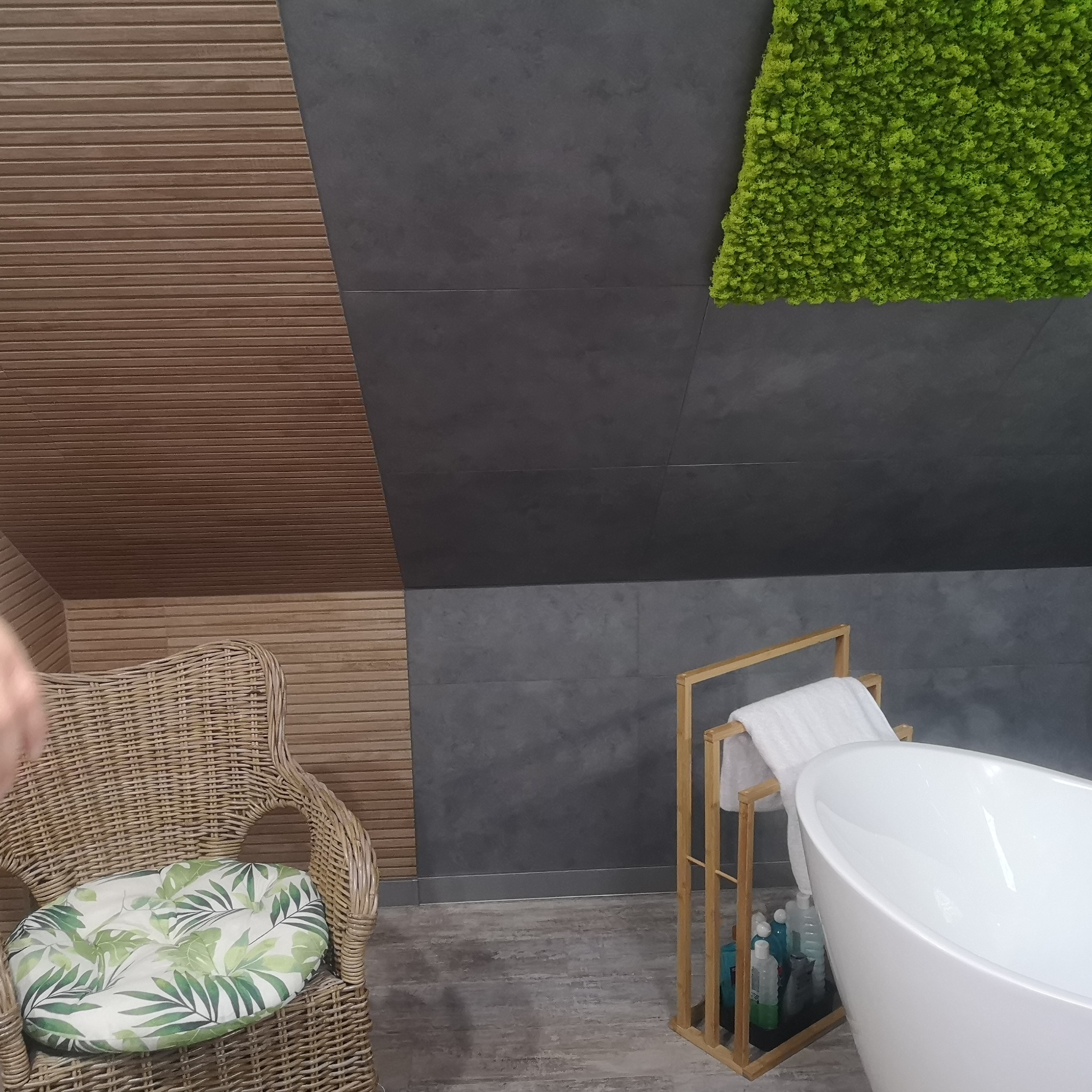Nowoczesna łazienka na poddaszu z drewnianą boazerią, szarą płytką imitującą beton, zielonym panelem z mchu, wiklinowym fotelem z poduszką w liście i wolnostojącą białą wanną z drewnianym...