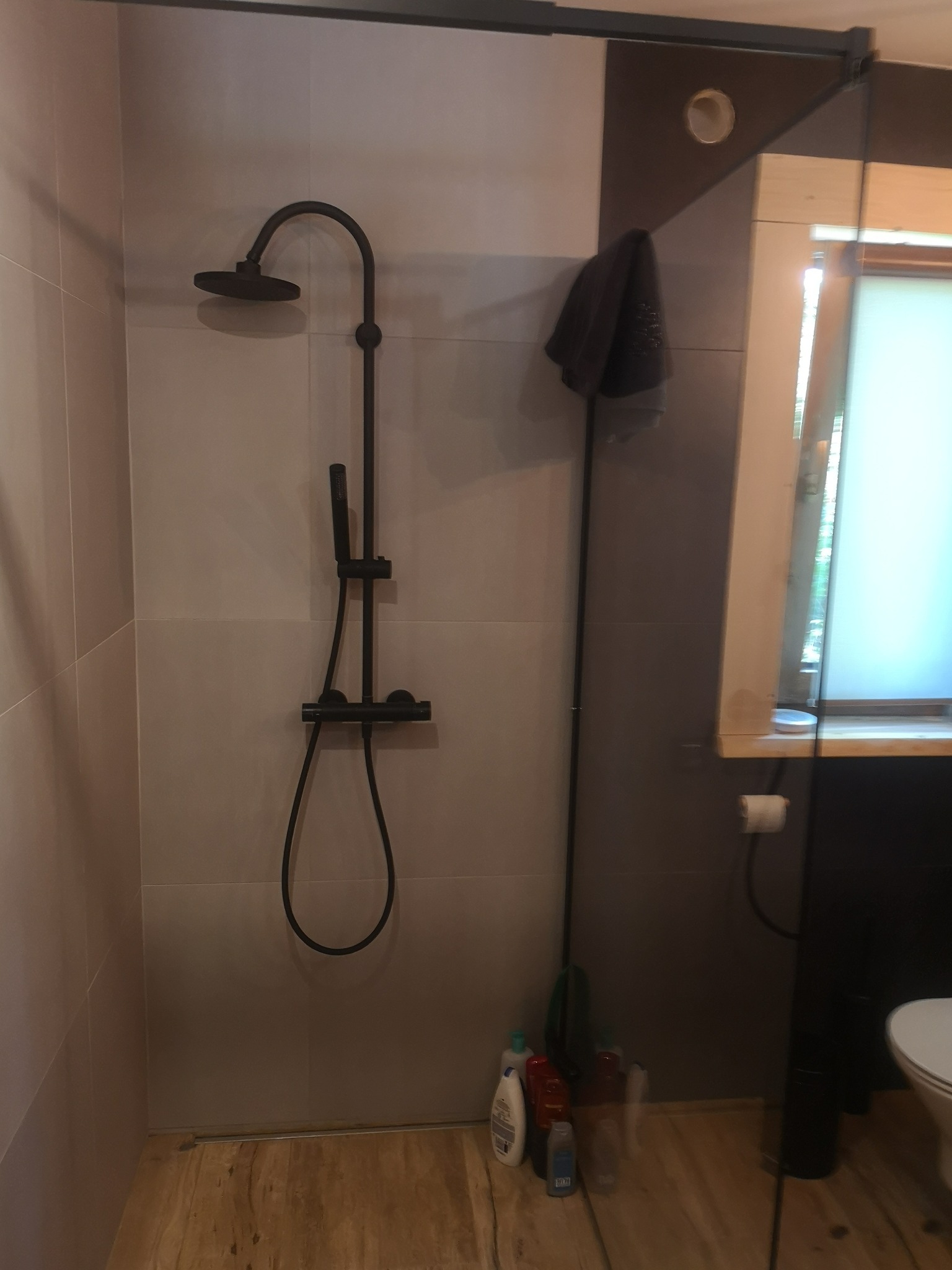 Nowoczesny prysznic z deszczownicą w kolorze czarnym, szare płytki ścienne, drewnopodobna podłoga, widoczne butelki z kosmetykami i ręcznik.