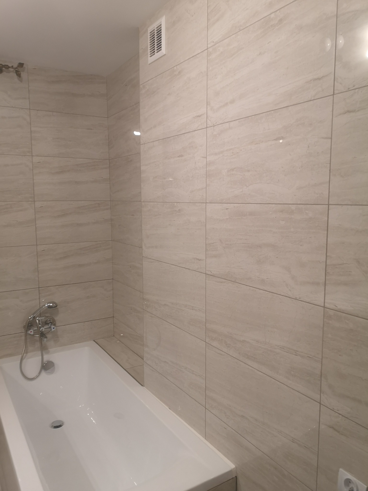 Biała wanna z chromowaną armaturą na tle ściany wyłożonej beżowymi płytkami imitującymi trawertyn, widoczny fragment wentylacji w górnej części ściany.