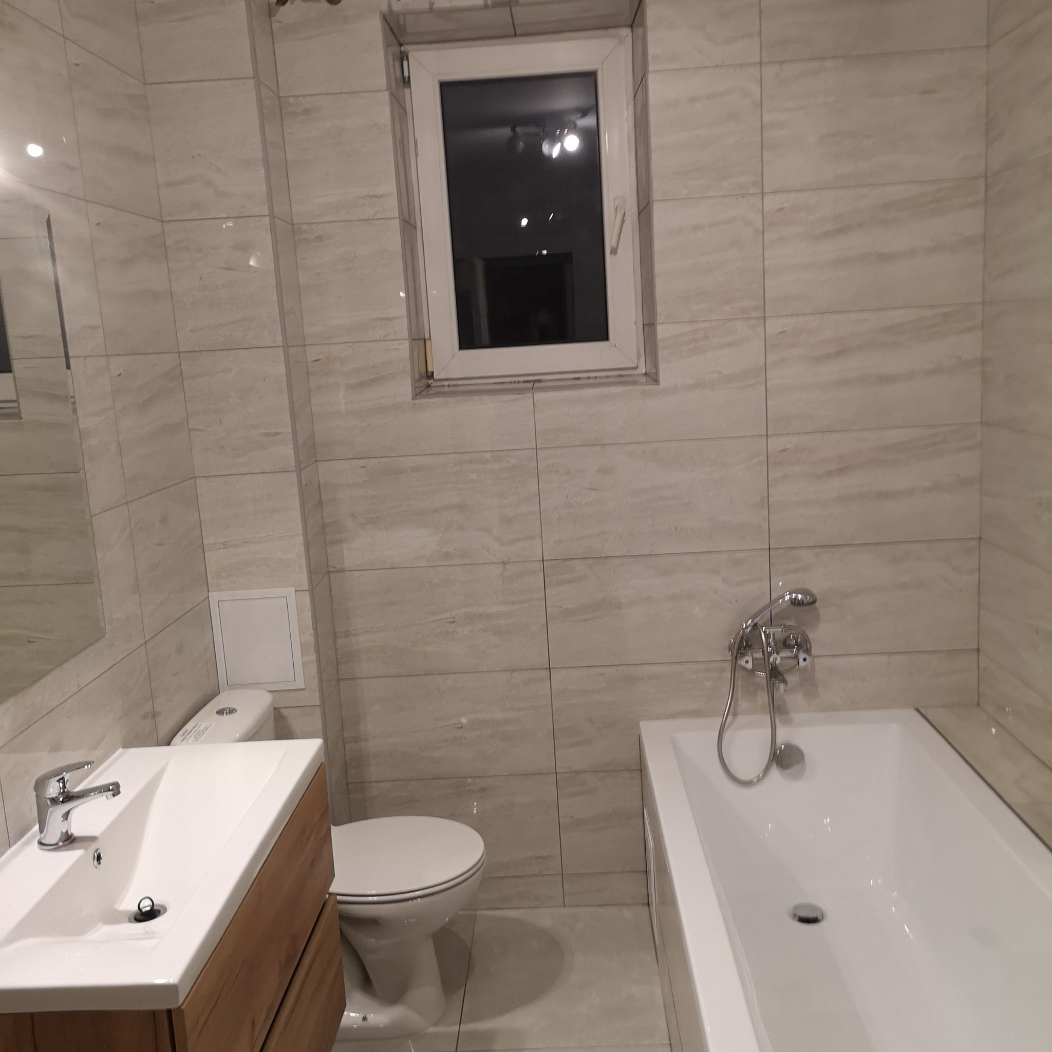 Jasna łazienka z beżowymi płytkami imitującymi marmur, białą wanną, umywalką z drewnianą szafką i toaletą w zabudowie.