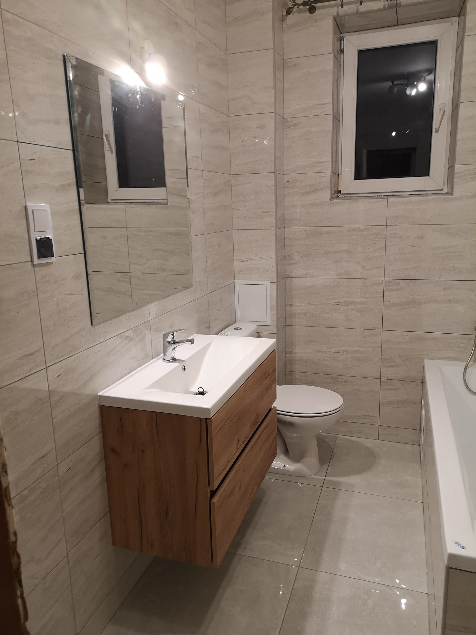 Nowoczesna łazienka z beżowymi płytkami imitującymi trawertyn, drewnianą szafką pod umywalką i dużym lustrem bez ramy, oświetlonym lampą nad lustrem.