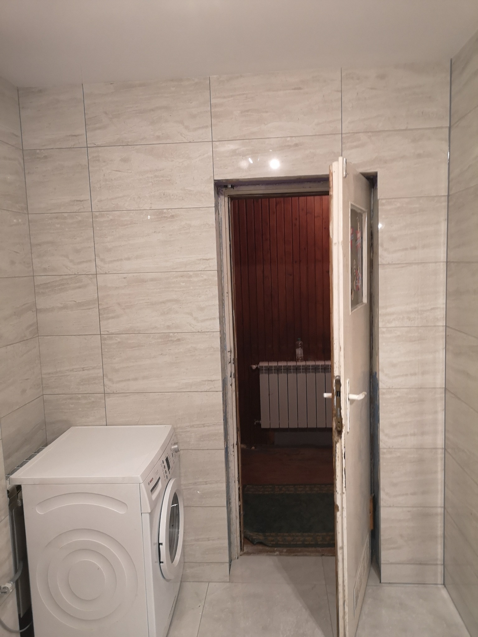 Wykończona łazienka z beżowymi płytkami imitującymi marmur, biała pralka po lewej stronie, otwarte drzwi do innego pomieszczenia z grzejnikiem i boazerią na ścianie.