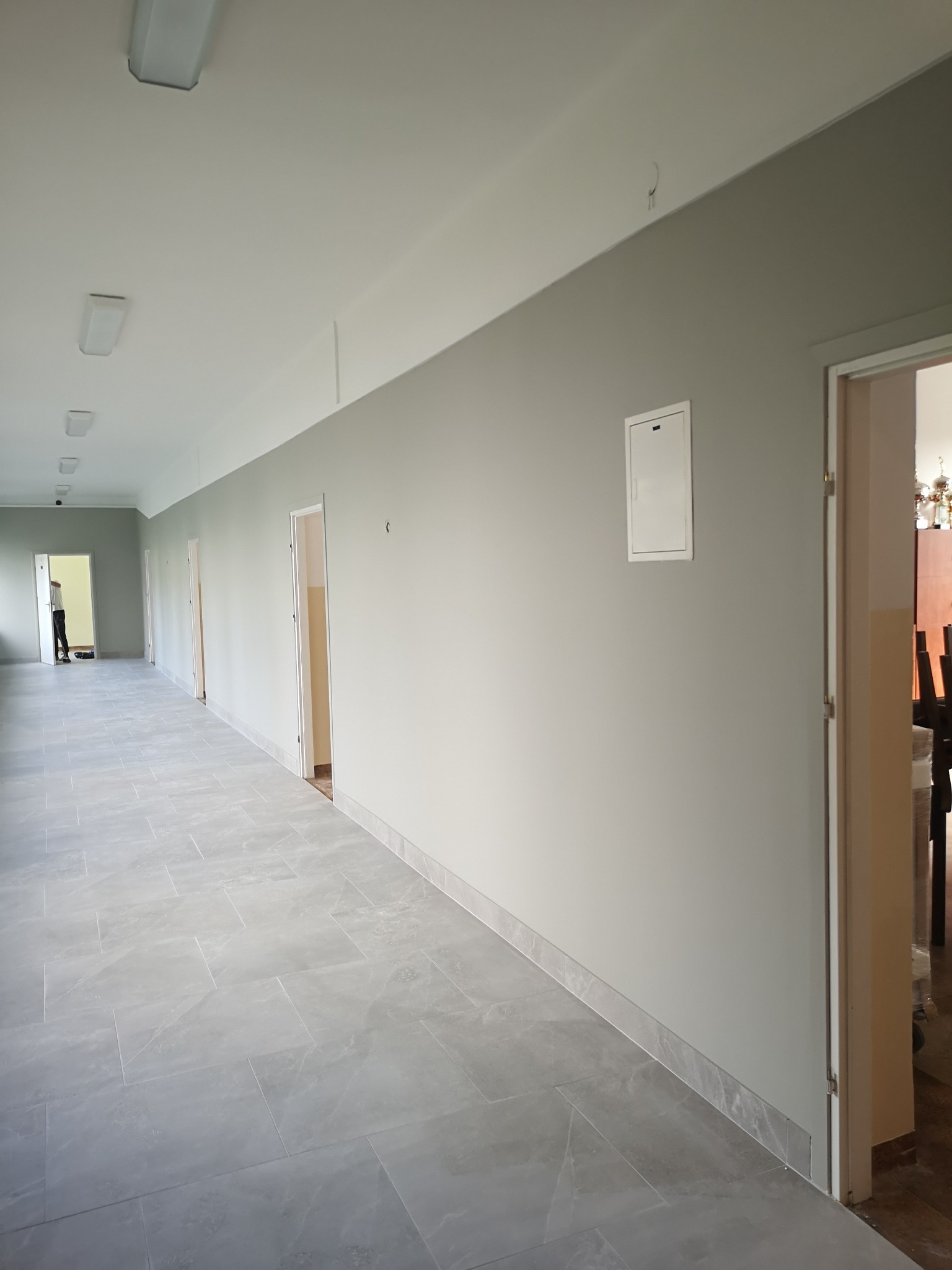 Długi korytarz w budynku biurowym z szarymi ścianami, szarą podłogą z płytek i białymi drzwiami, widoczne oświetlenie sufitowe i biała skrzynka na ścianie.