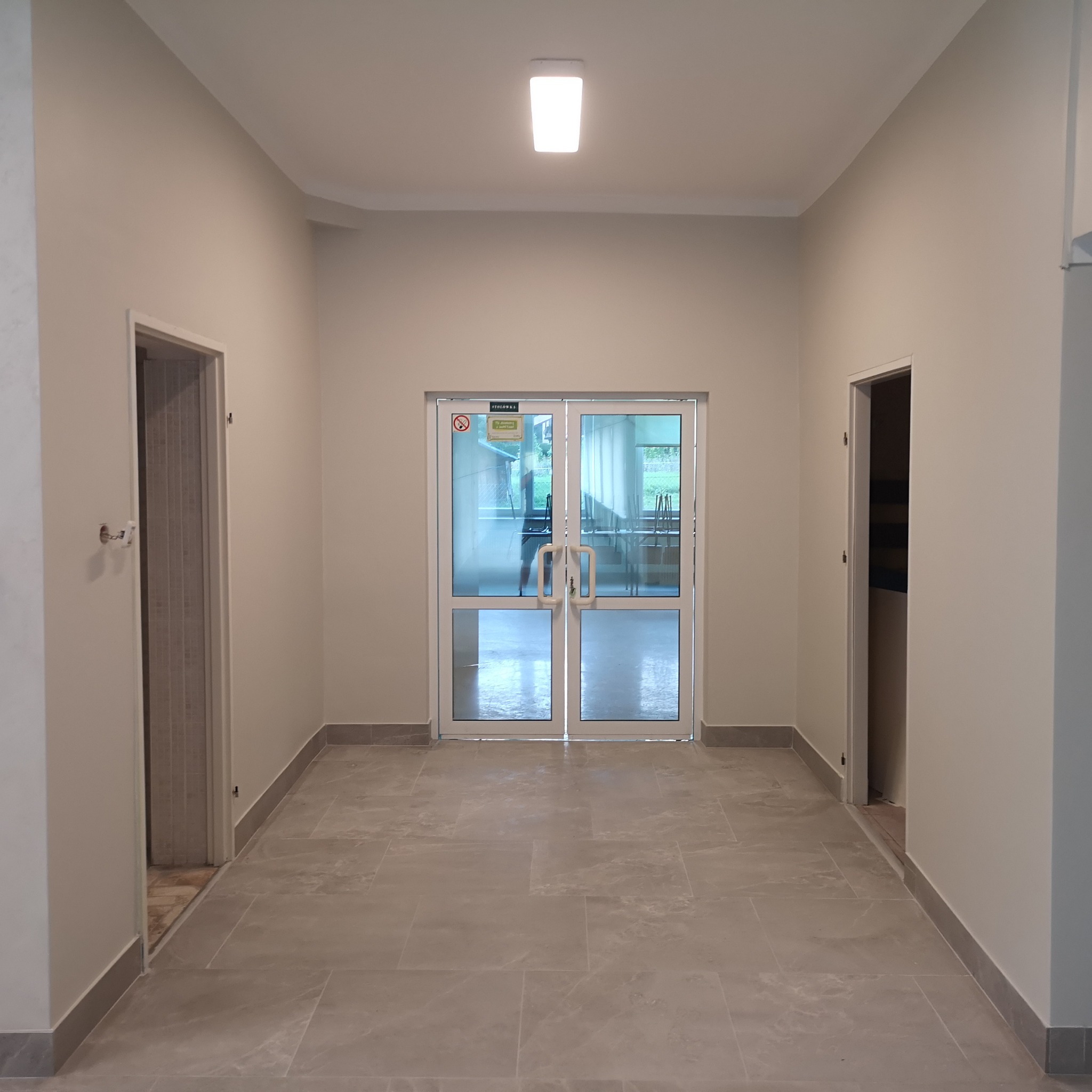 Korytarz biurowy z szarymi płytkami podłogowymi, świeżo pomalowanymi ścianami w odcieniu beżu i białymi drzwiami z szybami.