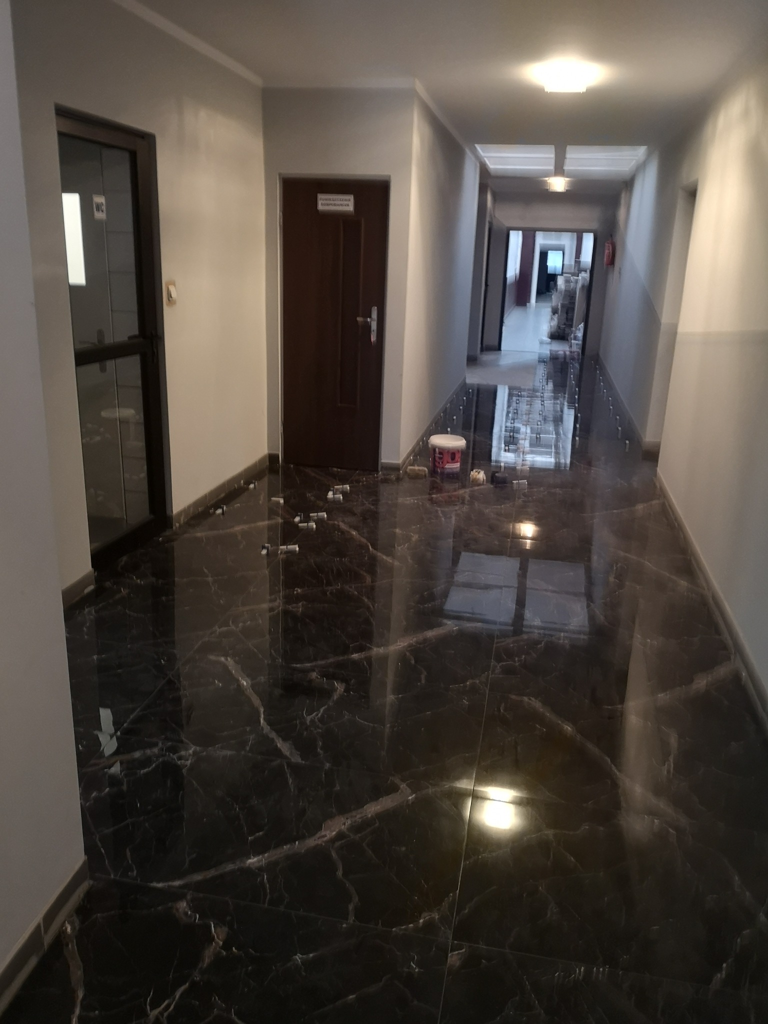 Położona, lśniąca, czarna glazura imitująca marmur w korytarzu w Komańczy, widoczne białe kliny dystansowe i wiaderko po farbie, sugerujące trwający remont.
