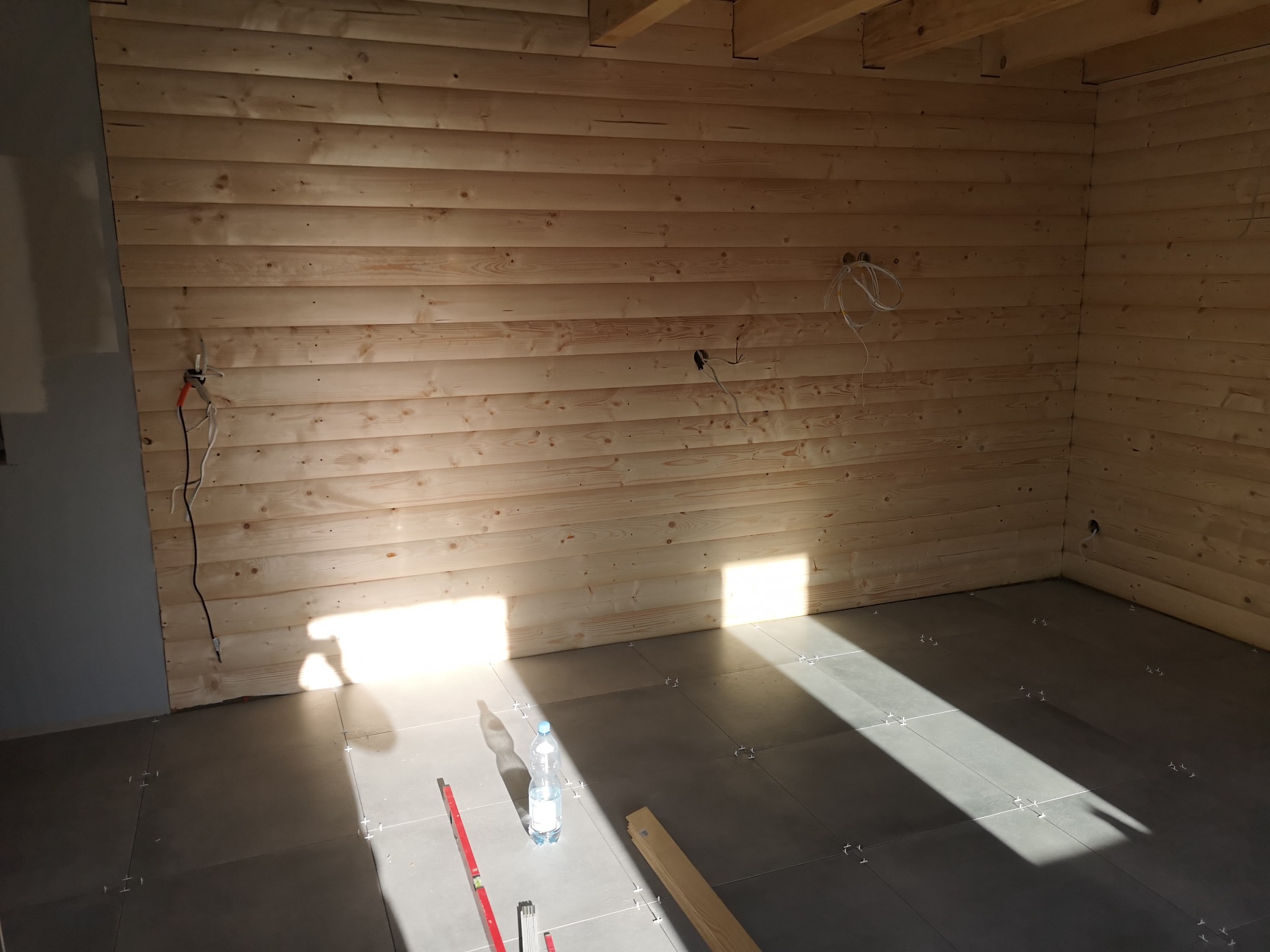 Wnętrze w trakcie prac: drewniana boazeria na ścianach, szare płytki podłogowe z krzyżykami dystansowymi, widoczne kable elektryczne, poziomica i butelka wody.