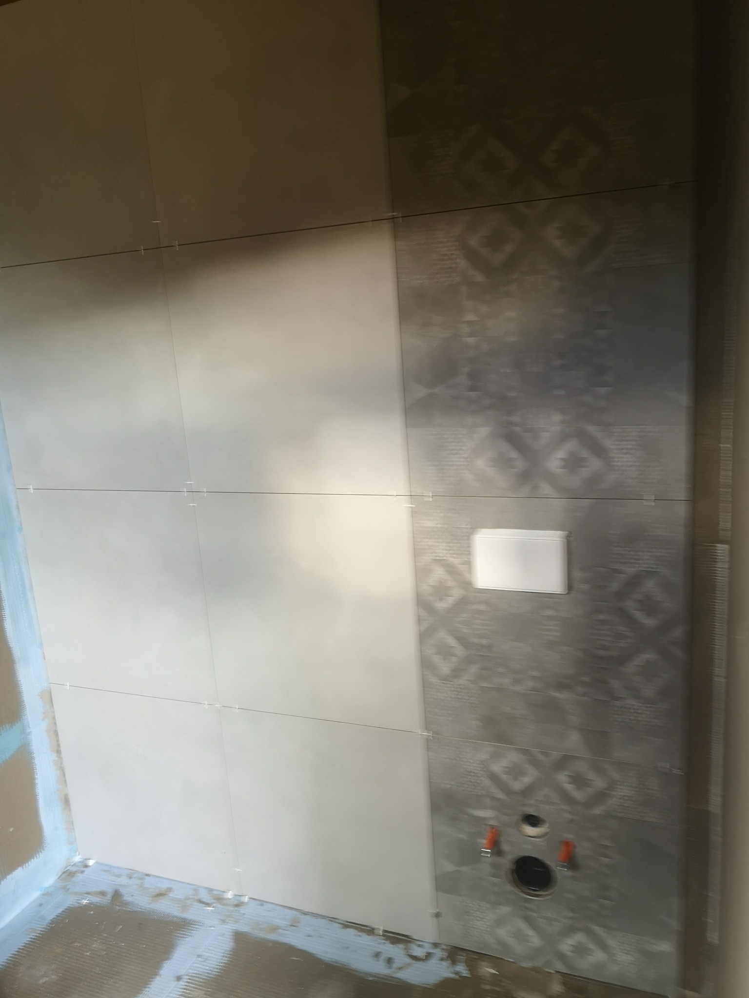 Ściana łazienki w trakcie układania płytek: jasnoszare kwadratowe płytki z jednej strony i ciemnoszare z wzorem z drugiej strony, widoczne małe krzyżyki dystansowe, instalacja podtynkowego WC...