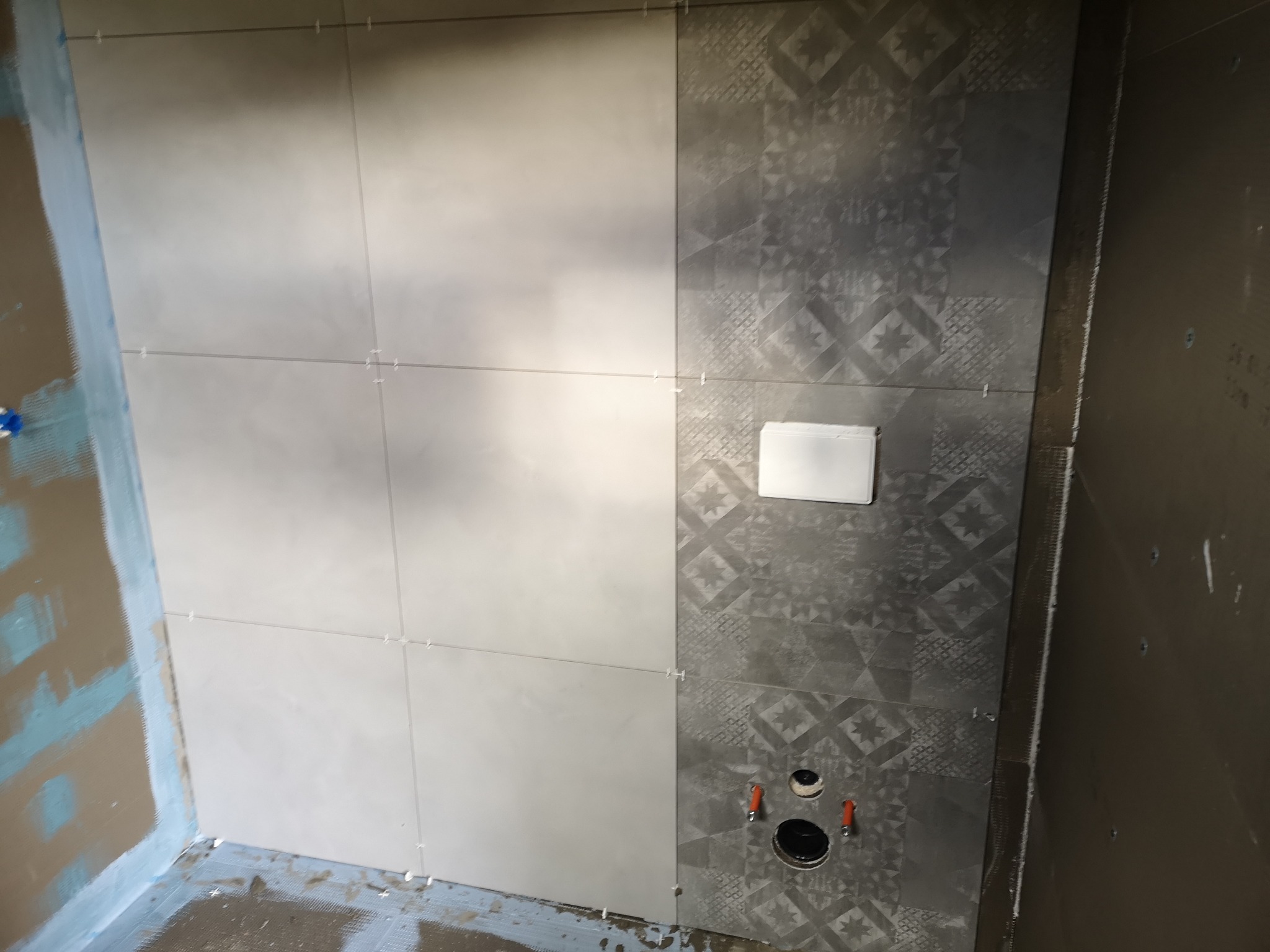 Układanie płytek w łazience: połączenie gładkich, jasnych płytek z dekoracyjnymi szarymi, widoczne krzyżyki dystansowe, przygotowane otwory na instalację sanitarną, ściana przygotowana pod płytki.