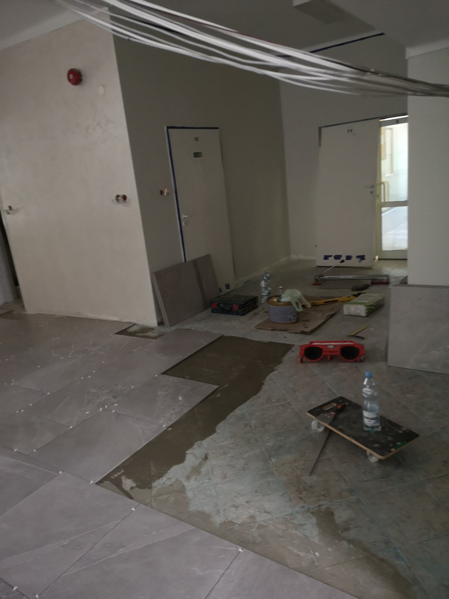 Układanie szarych płytek podłogowych z użyciem krzyżyków dystansowych, widoczne narzędzia i materiały budowlane, niedokończona podłoga w trakcie prac.