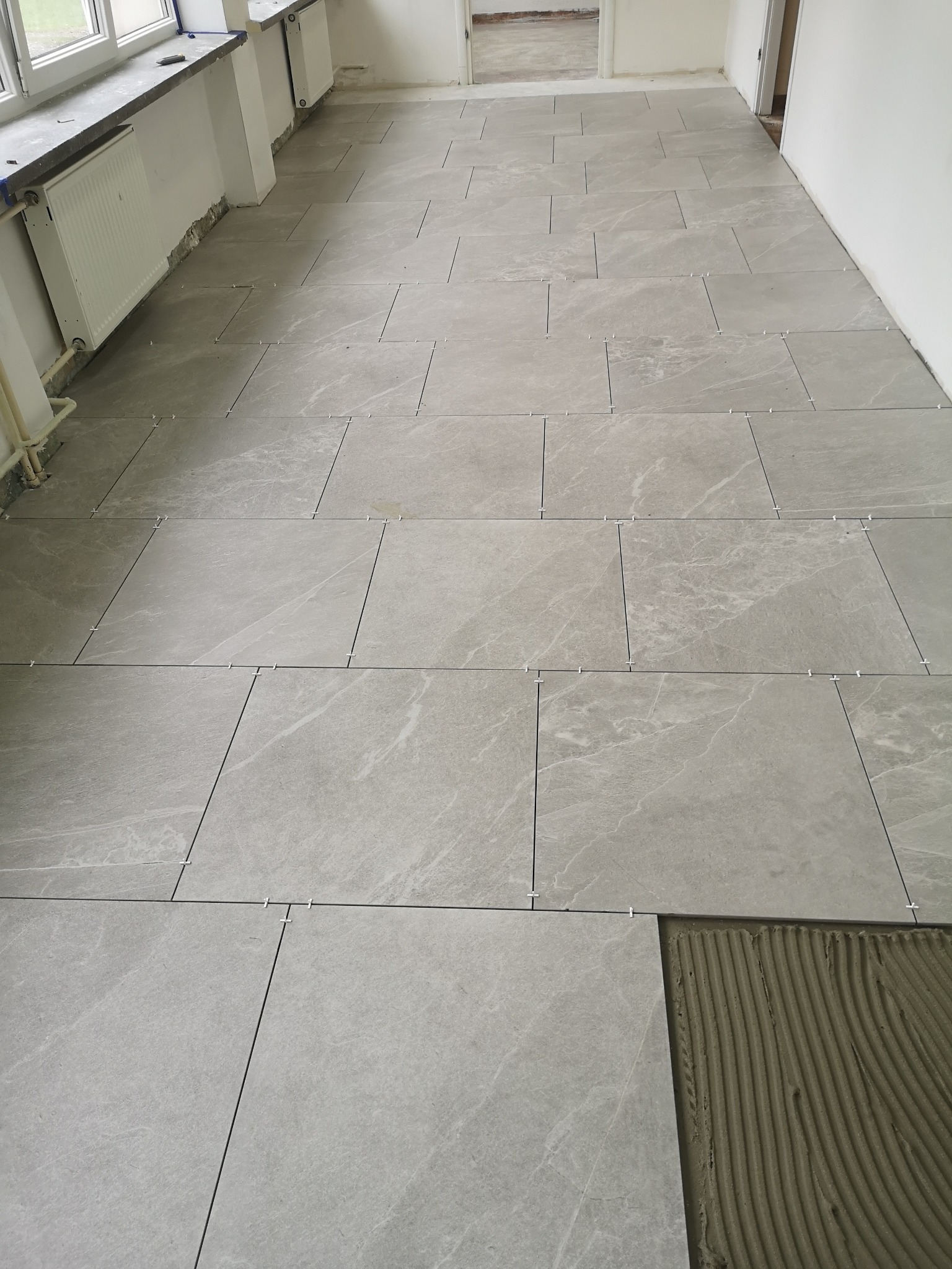 Układanie szarych płytek podłogowych w trakcie prac, widoczne krzyżyki dystansowe i warstwa kleju pod ostatnią płytką.