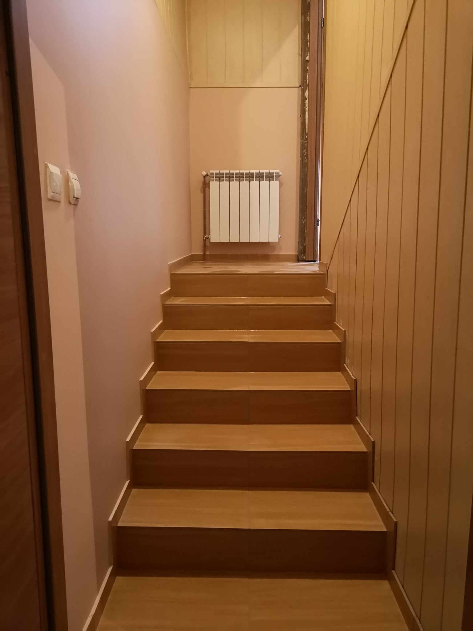 Schody wyłożone płytkami imitującymi drewno, prowadzące na wyższą kondygnację, ściana z boazerią w pionowe pasy, różowa ściana z grzejnikiem.