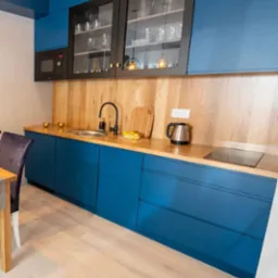 Niebieska kuchnia na wymiar z drewnianym blatem i czarnymi akcentami, przeszklone szafki wiszące z dekoracyjnym oświetleniem, nowoczesny design.