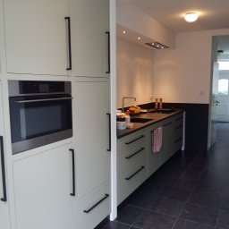 Bartosz Szyszka - Nowoczesna kuchnia z jasnymi szafkami z długimi, czarnymi uchwytami i wbudowanym piekarnikiem, z ciemnym blatem i czarną ścianą w tle, widok na przedpokój z białymi drzwiami wejściowymi.