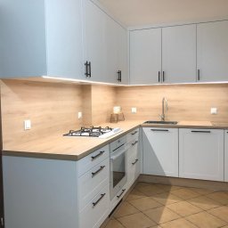 Bartosz Szyszka - Jasna kuchnia z białymi szafkami i drewnianym blatem, oświetlenie LED pod szafkami wiszącymi, czarne, minimalistyczne uchwyty, płyta gazowa na blacie.