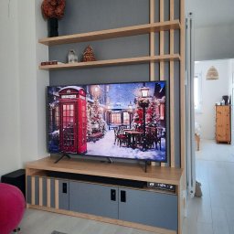 Bartosz Szyszka - Nowoczesna szafka RTV w odcieniach drewna i szarości, z widocznymi elementami dekoracyjnymi i półkami, na tle szarej ściany z drewnianymi listwami.