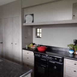 Bartosz Szyszka - Jasna kuchnia z czarnym piecem wolnostojącym, blatem z ciemnego kamienia i drewnianymi frontami szafek z czarnymi uchwytami. Na blacie stoi czerwony garnek, deska do krojenia i zioła w doniczkach.