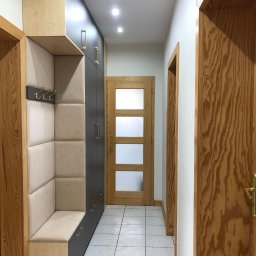 Bartosz Szyszka - Wnętrze przedpokoju z zabudową meblową: szafa w kolorze grafitowym z podłużnymi, złotymi uchwytami, obok siedzisko z tapicerowanym oparciem i wieszakami na ścianie. W tle widoczne drewniane drzwi...