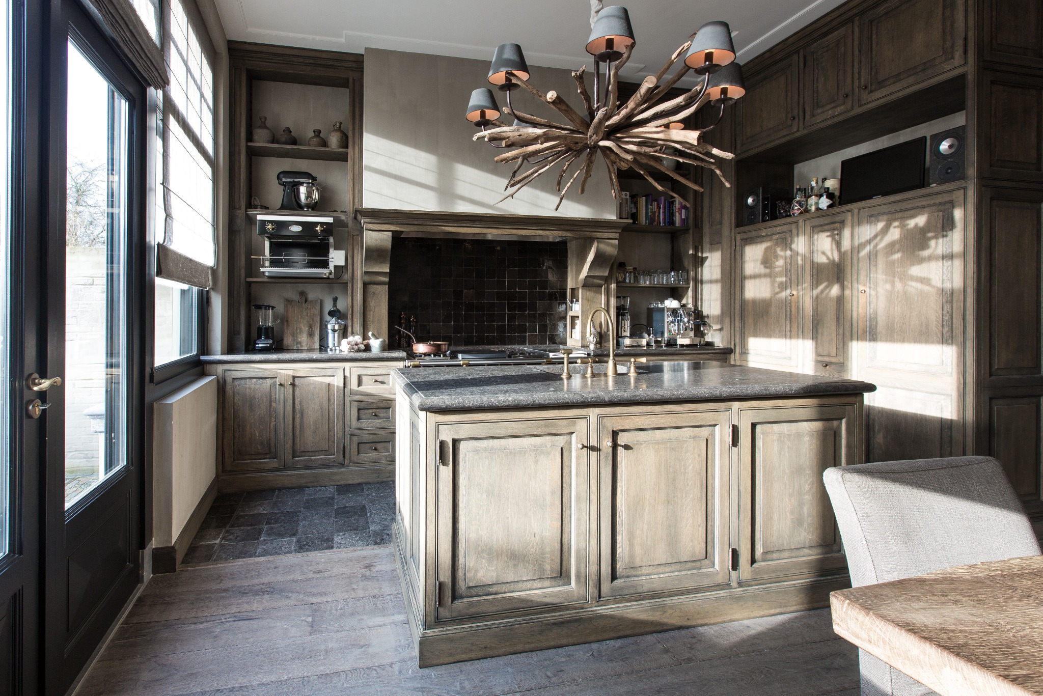 Przestronna kuchnia w odcieniach szarości z drewnianymi meblami, wyspą z kamiennym blatem, ozdobnym żyrandolem z gałęzi i dużym oknem wpuszczającym naturalne światło.