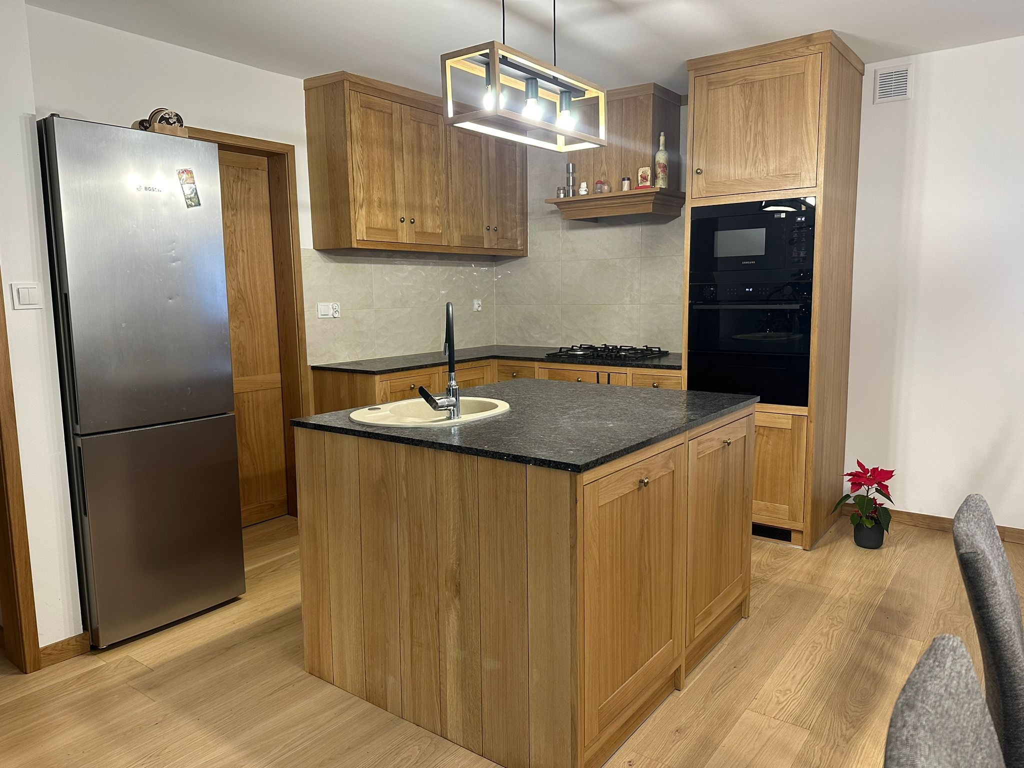 Jasna kuchnia z drewnianymi frontami, czarnym blatem i wyspą kuchenną z umywalką, oświetlona nowoczesną lampą wiszącą, z widoczną lodówką ze stali nierdzewnej i kwiatem doniczkowym.