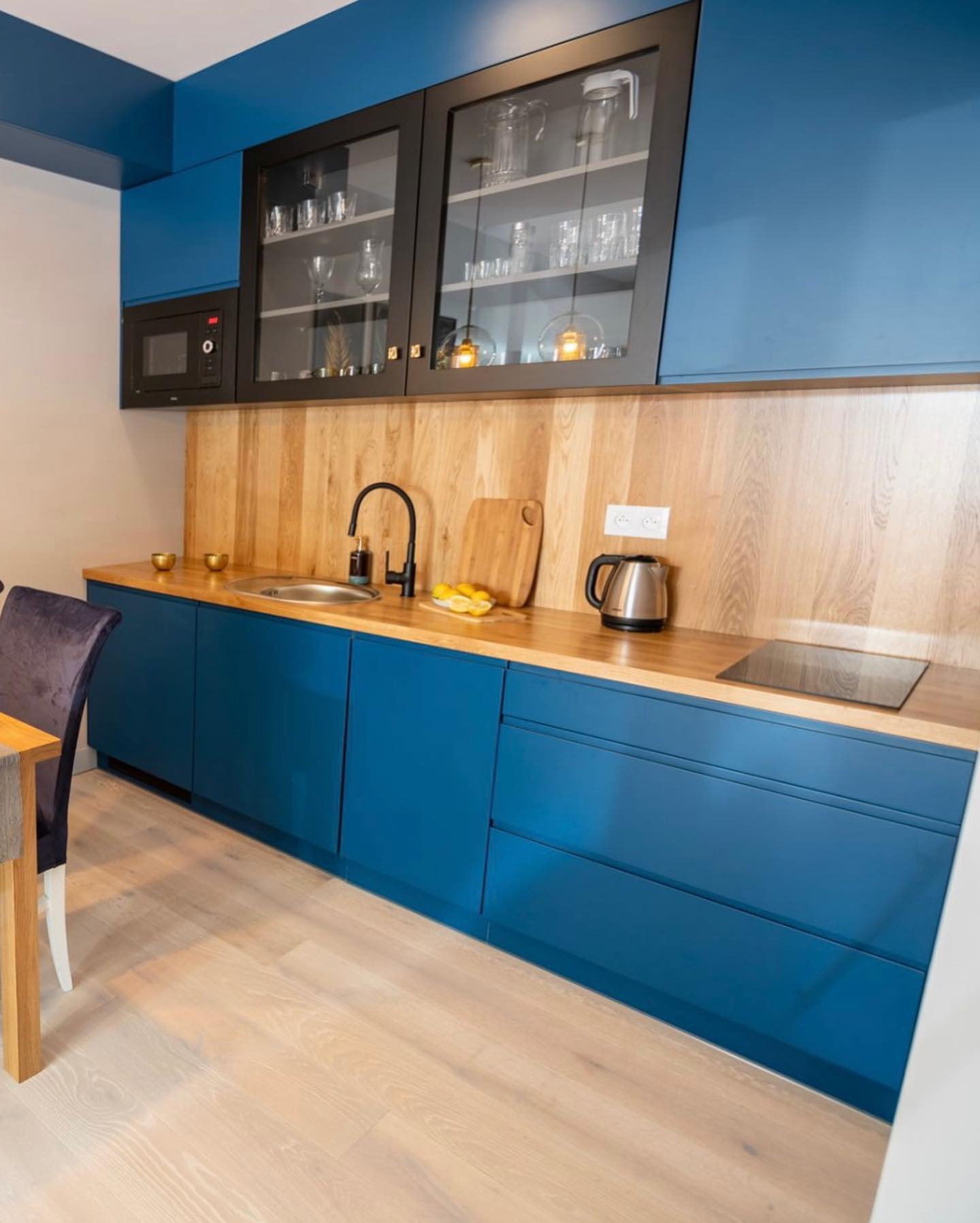 Niebieska kuchnia na wymiar z drewnianym blatem i czarnymi akcentami, przeszklone szafki wiszące z dekoracyjnym oświetleniem, nowoczesny design.