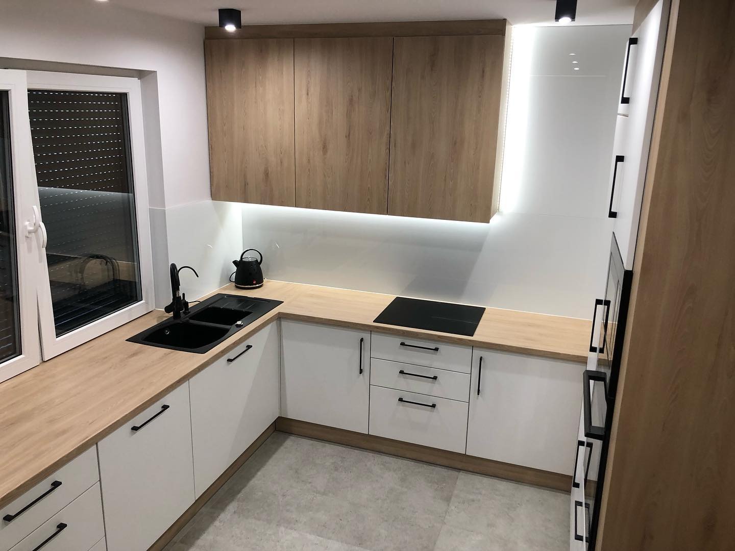 Nowoczesna, jasna kuchnia z białymi szafkami i drewnianymi blatami, czarny zlew i bateria przy oknie, czajnik na blacie, płyta indukcyjna, oświetlenie LED pod szafkami wiszącymi.