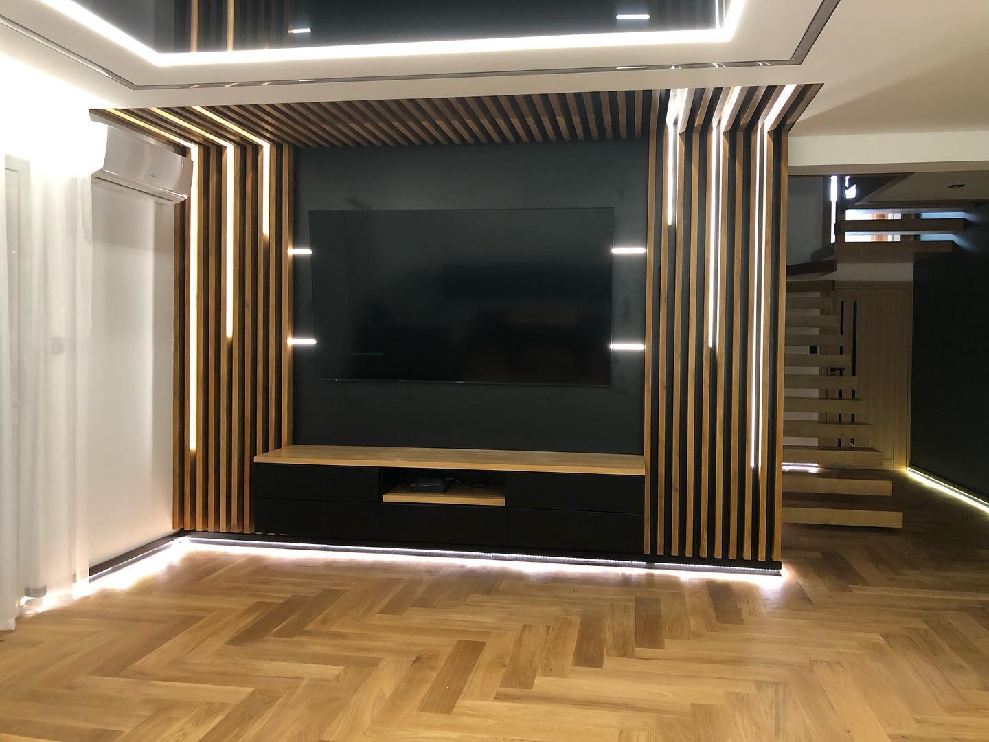 Nowoczesny salon z drewnianą zabudową telewizora z podświetleniem LED, parkietem w jodełkę i schodami w tle.